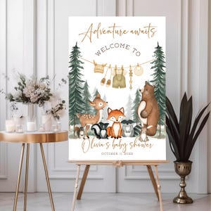 May include: A baby shower welcome sign on an easel. The sign features the text "Adventure awaits" and "Welcome to" in gold script. Below, illustrations of forest animals and baby clothes are displayed. The baby shower is for Olivia, October 11, 2026.
