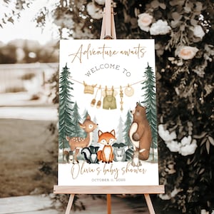 May include: A baby shower welcome sign on an easel. The sign features the text "Adventure awaits" and "Welcome to Olivia's baby shower" with the date. The sign has illustrations of forest animals and baby clothes hanging on a clothesline.