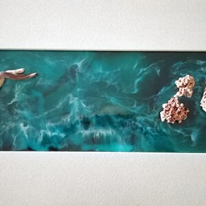 May include: Abstract ocean-themed artwork in a white frame. The piece features a teal and white resin base with a swirling pattern, resembling ocean waves.  It includes a driftwood-like sculpture and coral-like formations, creating a coastal aesthetic.