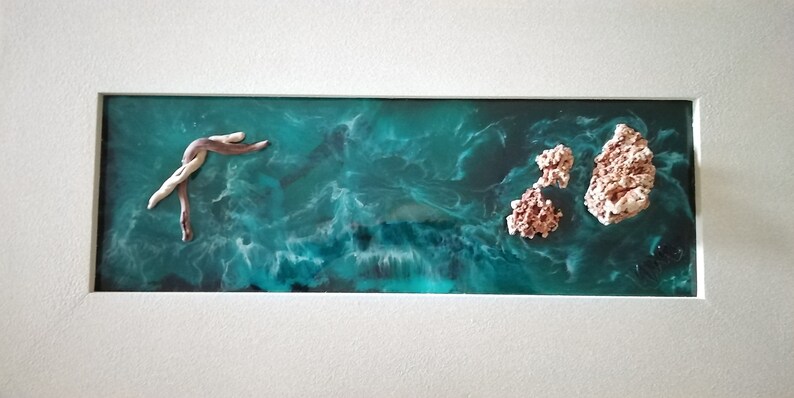 May include: Abstract ocean-themed artwork in a white frame. The piece features a teal and white resin base with a swirling pattern, resembling ocean waves.  It includes a driftwood-like sculpture and coral-like formations, creating a coastal aesthetic.
