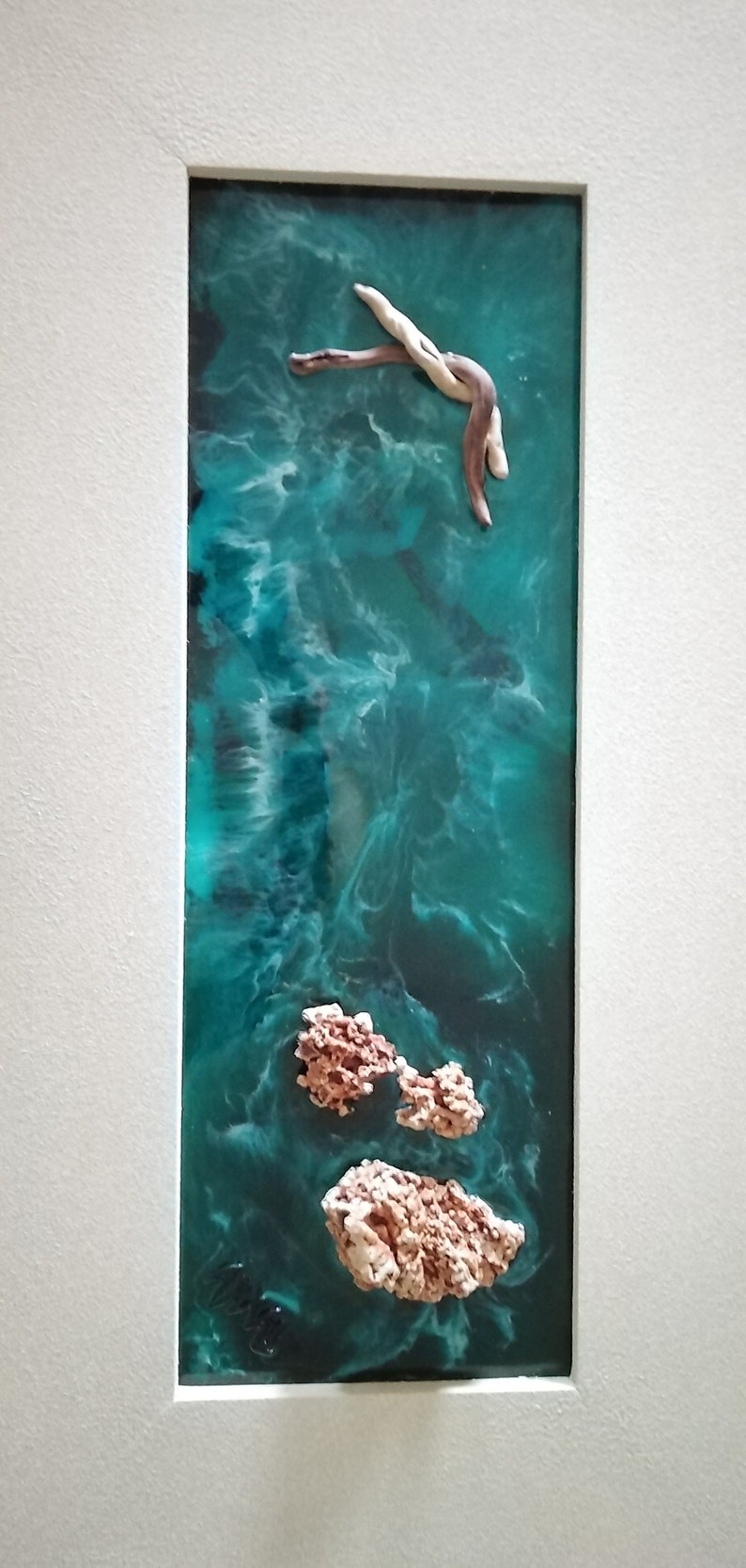 May include: Abstract resin art piece featuring a teal and turquoise ocean scene with a light-colored wooden element and coral-like formations. The artwork is framed in a white mat.