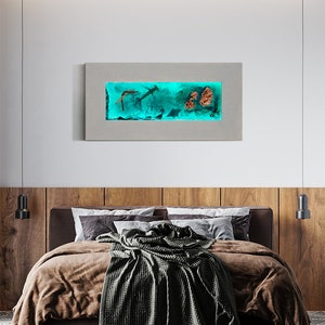 May include: A rectangular wall art piece featuring an underwater scene with sharks, coral, and other marine life. The artwork is encased in a gray frame and illuminated with a teal light, creating a vibrant display. The art hangs above a bed with brown bedding.