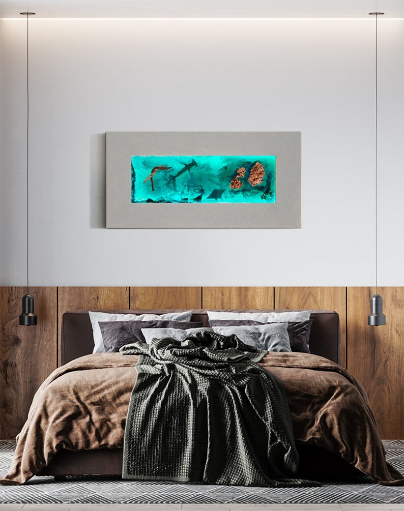 May include: A rectangular wall art piece featuring an underwater scene with sharks, coral, and other marine life. The artwork is encased in a gray frame and illuminated with a teal light, creating a vibrant display. The art hangs above a bed with brown bedding.