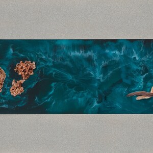 May include: Abstract ocean-themed artwork featuring a teal resin base with swirling patterns, resembling water. Two textured, coral-like formations and a piece of driftwood are embedded within the resin. The artwork is set within a rectangular, gray frame.