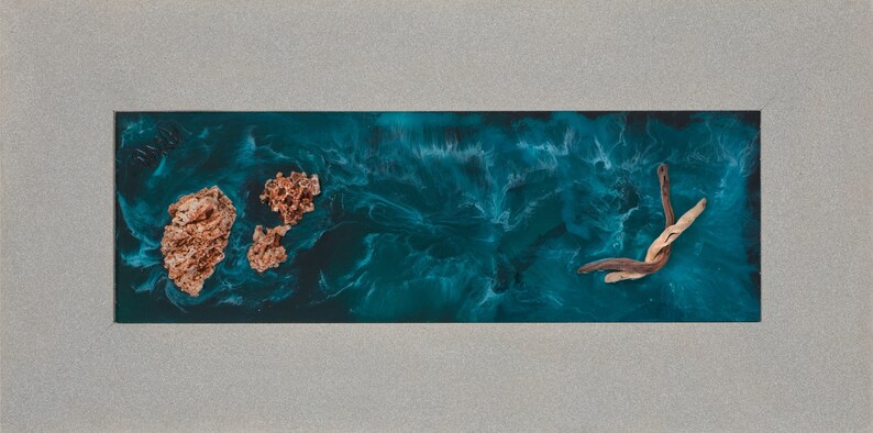 May include: Abstract ocean-themed artwork featuring a teal resin base with swirling patterns, resembling water. Two textured, coral-like formations and a piece of driftwood are embedded within the resin. The artwork is set within a rectangular, gray frame.