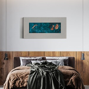 May include: A bedroom scene featuring a bed with brown bedding and a dark green blanket. Above the bed is a framed artwork with a teal background and gold accents. The headboard is made of wood panels, and the room has a modern design.