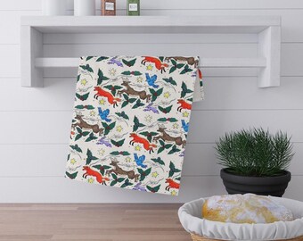 Op de afbeelding: Een crèmekleurige keukendoek met een herhalend patroon van rode vossen, bruine herten, blauwe vogels en groene hulstbladeren. Het ontwerp bevat ook gele sterren en paarse wolken. De handdoek hangt aan een witte plank.
