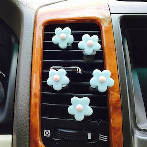 May include: Five light blue flower-shaped car air fresheners with pink centers are attached to a car's air vent.