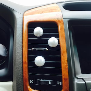 May include: Three white, pearl-like car vent air fresheners are attached to a black car vent. The vent is set in a wooden-look trim. The car's dashboard and controls are visible in the background.