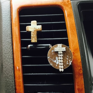 May include: Two car air vent decorations. One is a cross-shaped air freshener with a beige and brown speckled design. The other is a round, bronze-colored air freshener with a cross design made of rhinestones.