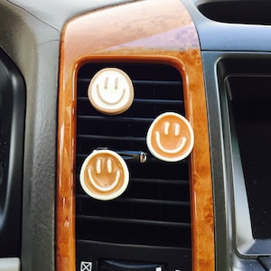 May include: Three smiley face car air fresheners, two are brown with white outlines and one is white with brown outlines, attached to a car's air vent.