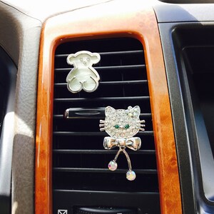 May include: Two decorative car vent clips. One is a silver teddy bear shape with a white centre. The other is a silver cat face with rhinestone accents, green eyes, and a bow. The clips are attached to a car vent.