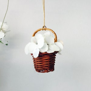 May include: A small brown wicker basket with a white handle is filled with white paper flowers. The basket is hanging from a string.