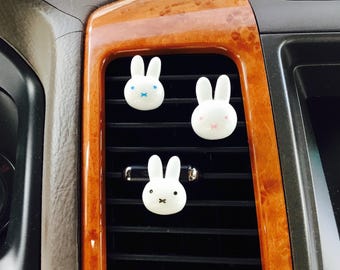 Pode incluir: Três ambientadores brancos para carro em forma de coelho com detalhes faciais pretos, azuis e rosa são exibidos em uma ventilação do carro. O interior do carro apresenta acabamento em grão de madeira. Os coelhos têm um design minimalista.