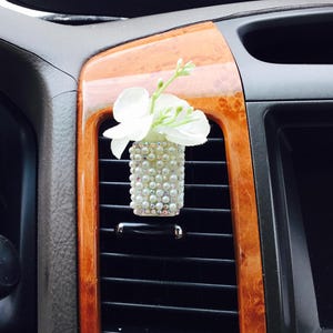 May include: White flower air freshener with pearl accents in a car air vent.