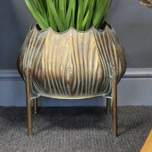 May include: A gold metal planter with a textured, ribbed design and a wide, rounded base. The planter is on a stand with three legs and is filled with green plants.