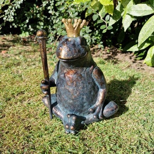 May include: A bronze frog statue wearing a gold crown and holding a staff. The frog is seated on green grass, with a natural background of foliage. The statue has a weathered, antique appearance.