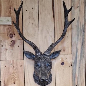 May include: A dark grey deer head wall mount with large antlers. The deer head is mounted on a wooden plank wall. The antlers are a darker shade of grey than the head.