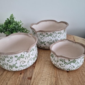 May include: A trio of ceramic plant pots with a scalloped edge. The pots are white with a green floral design and a brown rim. They are displayed on a wooden surface.