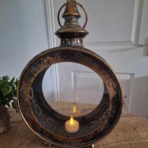 May include: An antique-style lantern with a circular design and a distressed bronze finish. It features a clear glass panel and an LED candle inside, providing a warm glow. The lantern is designed for indoor use and is a decorative item.
