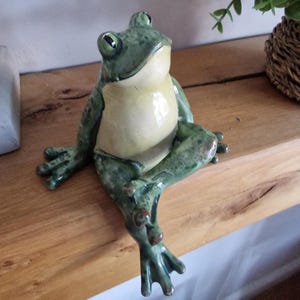 May include: A ceramic frog ornament in green and cream. The frog is depicted sitting with its legs crossed. The figurine has open eyes and is positioned on a wooden shelf.
