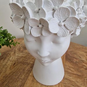 May include: A white ceramic head-shaped planter adorned with a floral crown. The face features closed eyes and a calm expression. The planter sits on a wooden surface, with a small green plant in the background.