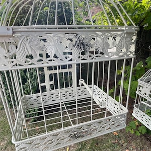 May include: Off-white decorative birdcage with a domed top and vertical bars. The cage features decorative grape and leaf accents along the top and bottom edges. The cage is open, with a small door and a latch.