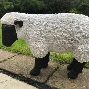 May include: White sheep statue with a black face and legs. The sheep is made of a textured material that resembles wool.