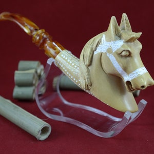 May include: A carved tobacco pipe with a horse head design. The pipe features a tan-coloured horse head with white accents and a brown stem. The pipe is displayed on a clear stand, with several bamboo tubes nearby.