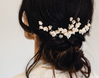 May include: A woman with dark brown hair styled in a low bun with a delicate floral hair comb. The comb features small white flowers and gold accents.