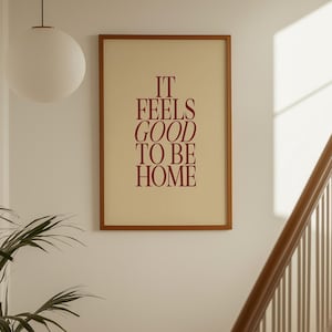 May include: A framed print with the text "IT FEELS GOOD TO BE HOME" in burgundy lettering. The print has a cream-coloured background and a brown frame. The image is hanging on a white wall near a staircase.