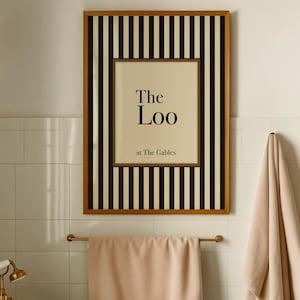 May include: A framed print reading "The Loo at The Gables" is displayed on a white tiled wall. The frame features a black and white striped border. A peach-coloured towel hangs on a gold-coloured rail, with another towel on a hook.