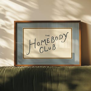 May include: A framed print with the text "THE HOMEBODY CLUB" in a handwritten style. The artwork is set against a light beige background, framed in a wooden frame with a blue border. The print rests on a green velvet sofa.
