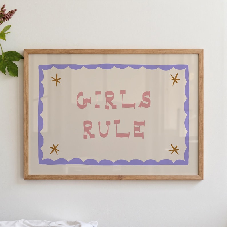 May include: A framed print with the words "GIRLS RULE" in pink letters. The text is centered on a cream-colored background, framed by a scalloped purple border and accented with gold star shapes in each corner. The frame is made of light brown wood.