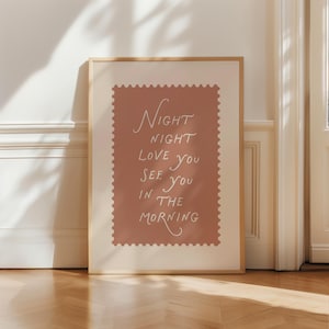May include: A framed art print featuring the words "Night Night Love You See You In The Morning" in white script on a dusty rose background. The print has a decorative border and is set in a light wood frame.
