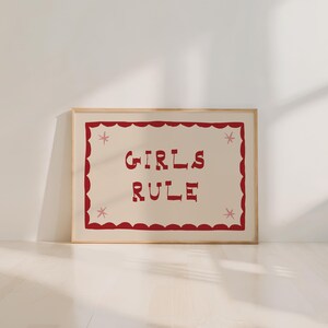May include: A framed print with the words "GIRLS RULE" in red, stylized lettering. The text is centered on a cream-colored background, framed by a red scalloped border with pink star accents. The frame is a light wood color.