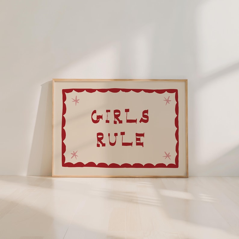 May include: A framed print with the words "GIRLS RULE" in red, stylized lettering. The text is centered on a cream-colored background, framed by a red scalloped border with pink star accents. The frame is a light wood color.