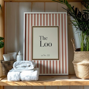 Könnte beinhalten: Ein gerahmter Druck mit vertikalen Streifen in Weiß und Staubrosa, der den Text "The Loo at No. 22" zeigt. Der Druck befindet sich auf einem Holzregal mit gefalteten weißen Handtüchern, Toilettenartikeln und einem geflochtenen Korb mit einer Topfpflanze.