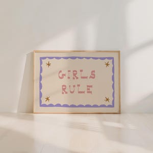 May include: A framed art print with the words "GIRLS RULE" in pink letters on a cream background. The print has a purple scalloped border and gold star accents. The frame is a light wood color.