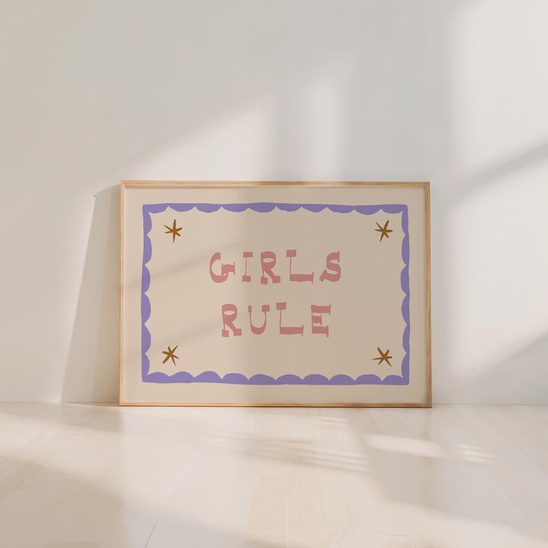 May include: A framed art print with the words "GIRLS RULE" in pink letters on a cream background. The print has a purple scalloped border and gold star accents. The frame is a light wood color.