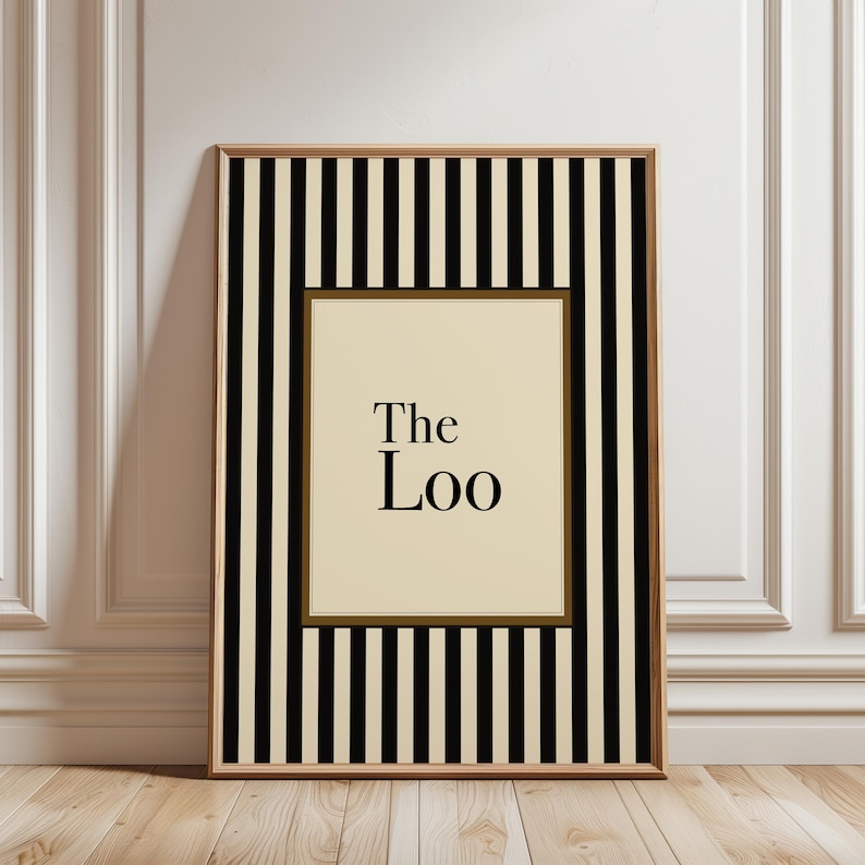 May include: A framed art print with a black and cream striped border. The inner frame is a light cream color with the words "The Loo" in black, centered. The frame is a light brown color.