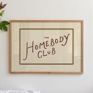 May include: Framed art print with the text "THE HOMEBODY CLUB" in a handwritten style. The text is in a dark red color against a cream background, framed by a thin brown border and a light wood frame. The artwork is displayed on a white wall.