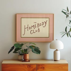 May include: A framed print with the text "The Homebody Club" in a handwritten style. The frame is a light pink with a gold border, and the print is displayed on a wooden cabinet with plants and a white lamp.