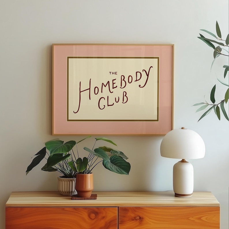 May include: A framed print with the text "The Homebody Club" in a handwritten style. The frame is a light pink with a gold border, and the print is displayed on a wooden cabinet with plants and a white lamp.