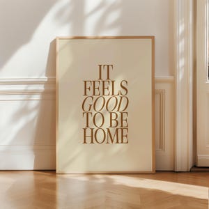 May include: A framed art print with the text "IT FEELS GOOD TO BE HOME" in a warm brown color. The artwork has a cream-colored background and is encased in a light brown wooden frame. The print is displayed against a white wall and a wooden floor.