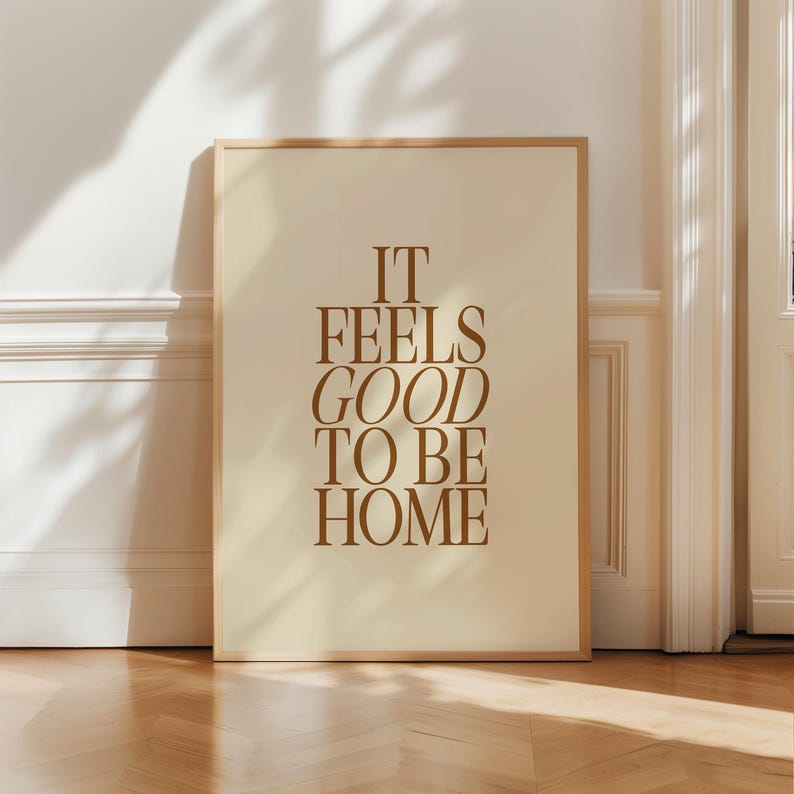 May include: A framed art print with the text "IT FEELS GOOD TO BE HOME" in a warm brown color. The artwork has a cream-colored background and is encased in a light brown wooden frame. The print is displayed against a white wall and a wooden floor.
