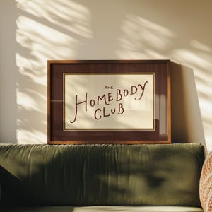 May include: A framed print with the text "THE HOMEBODY CLUB" in a handwritten style. The frame is a dark brown color, and the print has a cream-colored background. The print is displayed above a green velvet sofa.