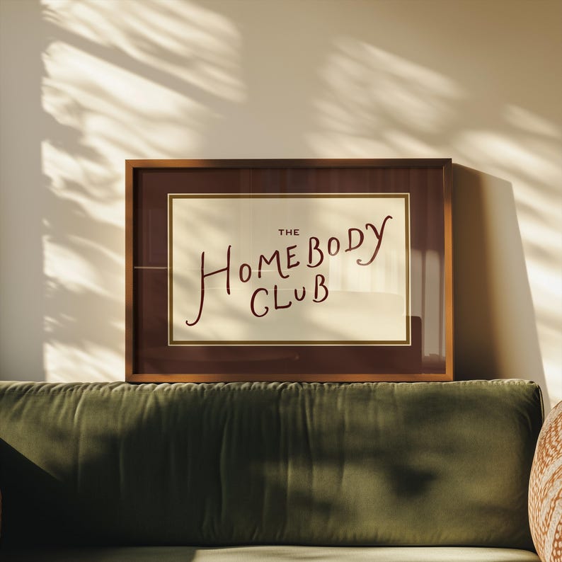 May include: A framed print with the text "THE HOMEBODY CLUB" in a handwritten style. The frame is a dark brown color, and the print has a cream-colored background. The print is displayed above a green velvet sofa.