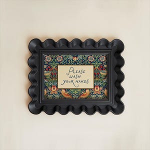 May include: A black, scalloped-edge picture frame with a floral patterned background and a cream-coloured sign that reads "Please Wash Your Hands" in elegant script. The frame is mounted on a light beige wall.