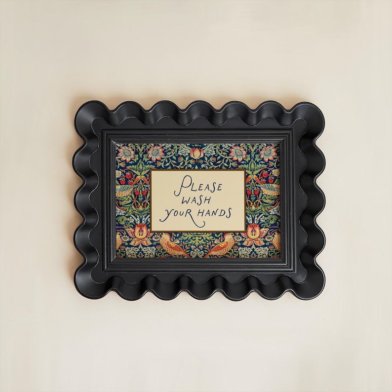 May include: A black, scalloped-edge picture frame with a floral patterned background and a cream-coloured sign that reads "Please Wash Your Hands" in elegant script. The frame is mounted on a light beige wall.